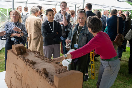 Sand sculptor Annie talking to guests about her sand sculpture of the new cancer centre at Great Ormond Street Hospital's event to celebrate the start of construction work. Marquee in park with guests standing and talking.