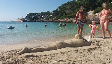 Full sized sand sculpture of a man reading a book on a towel on a beach.  In the background is a beautiful blue sea.  People are swimming and looking at the sculpture. The beach is in Cala D'Or, Majorca