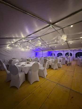 Innenansicht eines Partyzelts Berlin mit weißen Rundtischen, Stuhlhussen, Lichterketten und lila Ambientebeleuchtung – ideal für Hochzeiten und Geburtstagsfeiern