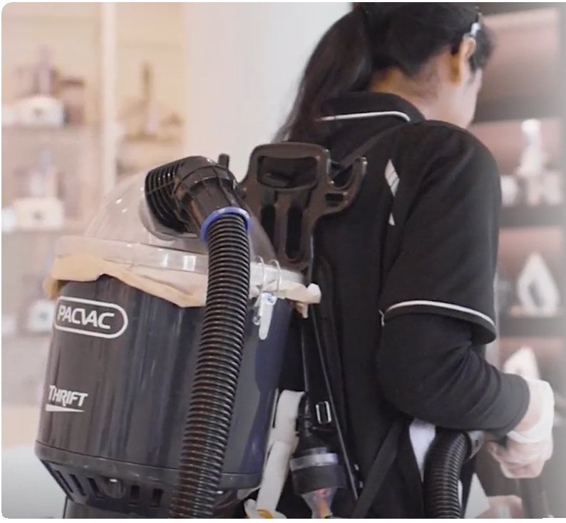 Person cleaning a room with a Pacvac Thrift backpack vacuum. They're wearing a black uniform with white accents. Shelves in the background.