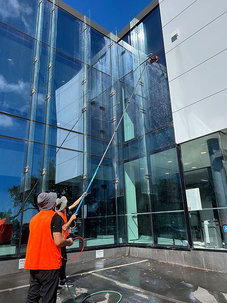 Two Men in High Visibility Vests use telescoping window cleaning machine on large glass panes.