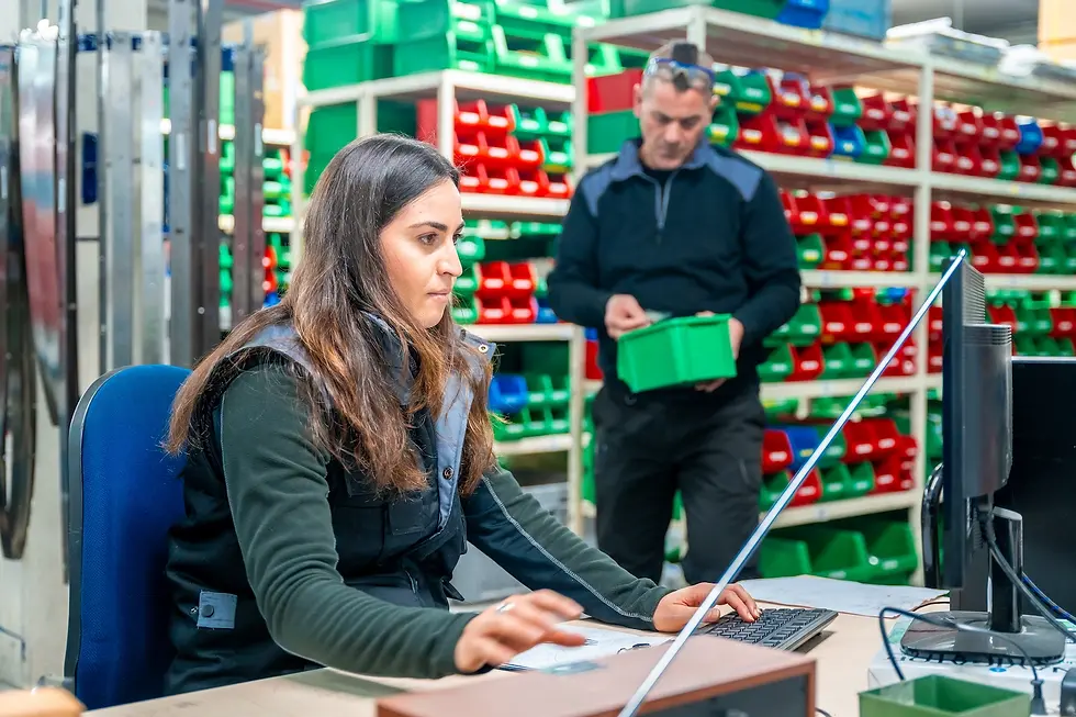 Woman working on a computer in a stockroom with colourful shelves. Man in background handles a green container.
