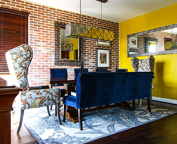 A vibrant dining room featuring an exposed red brick accent wall, a bold yellow statement wall, and rich blue upholstered seating. The room is adorned with a geometric pendant light, framed abstract art, and patterned wingback chairs with intricate bird and floral designs. A large silver-framed mirror reflects the space's eclectic style, while a blue and gray patterned rug anchors the wooden dining table and chairs.