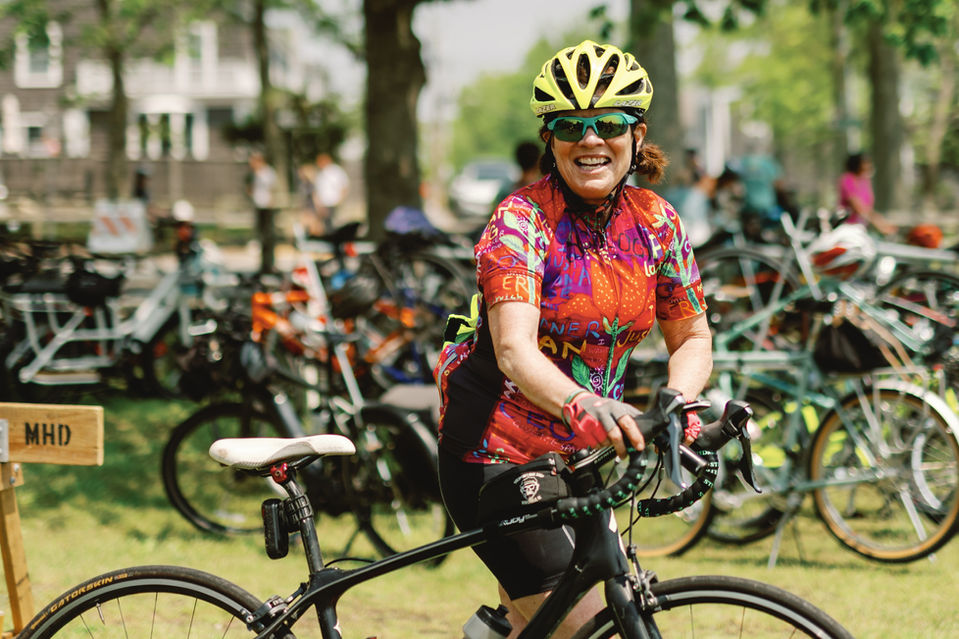 Woman in an orange rider shirt walking her bike into the park.