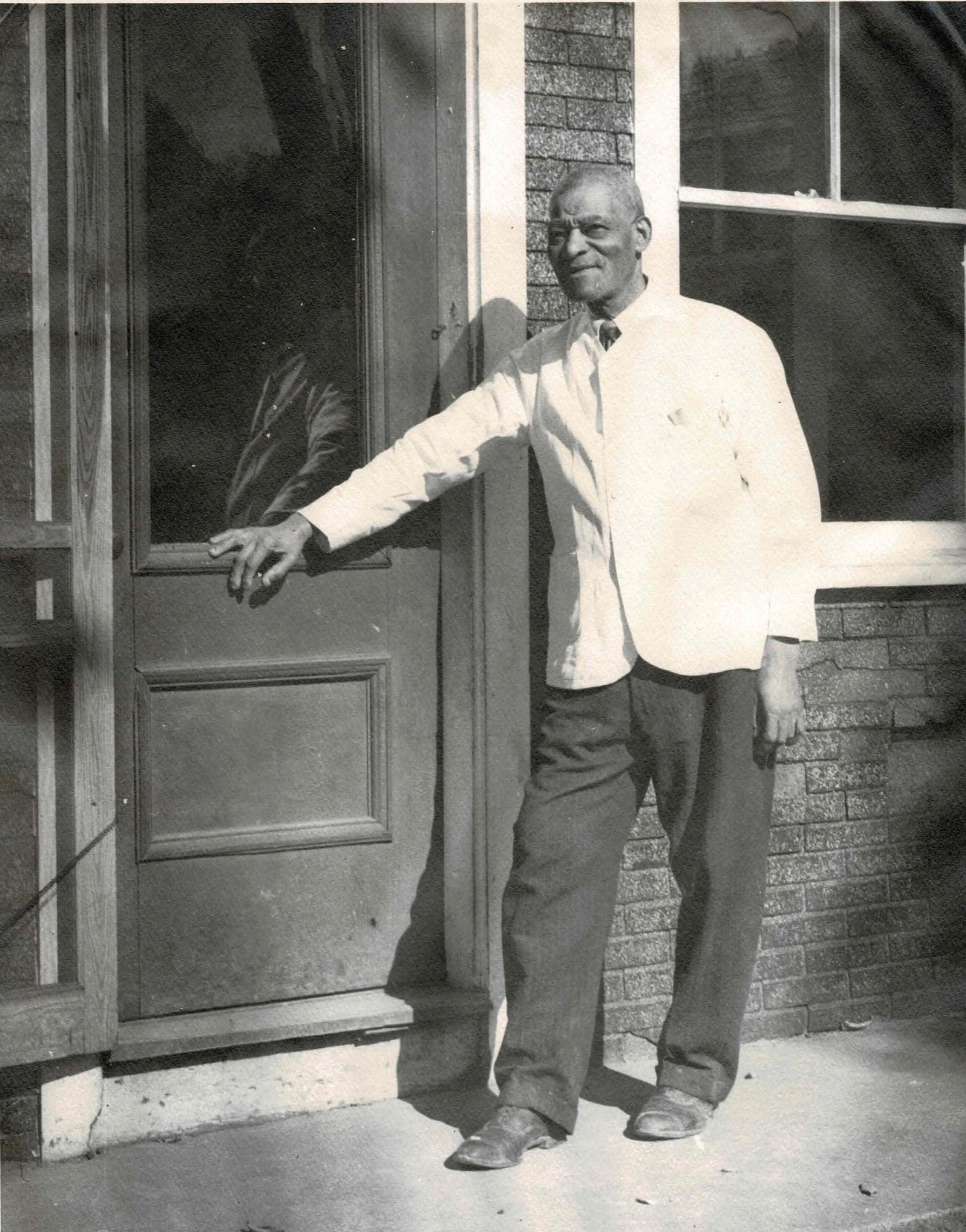 Black and white photo from the 1950's showing a black man smiling in a white coat outside of a barber shop