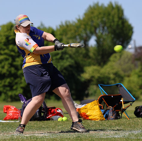 Person mid-swing hitting a softball, they are wearing a Raiders game shirt and have a rainbow cap on.