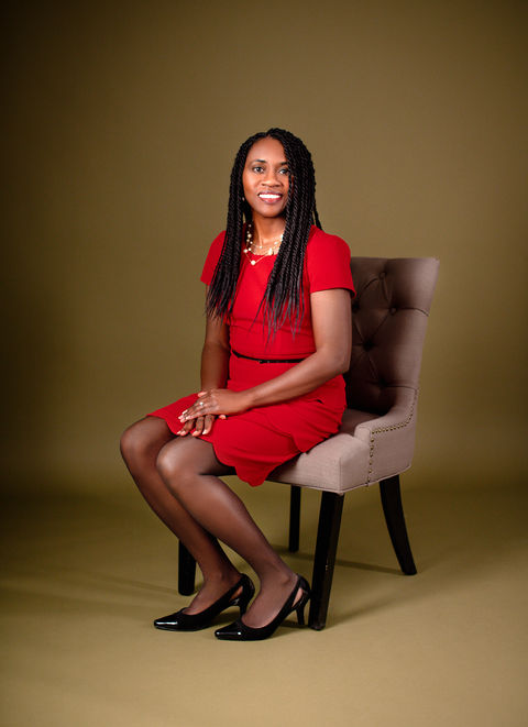 Gorgeous woman sits elegantly on a soft grey upholstered chair, posing for an individual portrait in front of a rich olive backdrop.