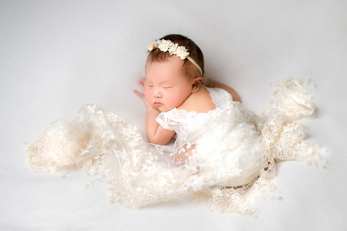 sleeping baby girl in white outfit lays on a plush white backdrop, draped in a cream lace blanket.