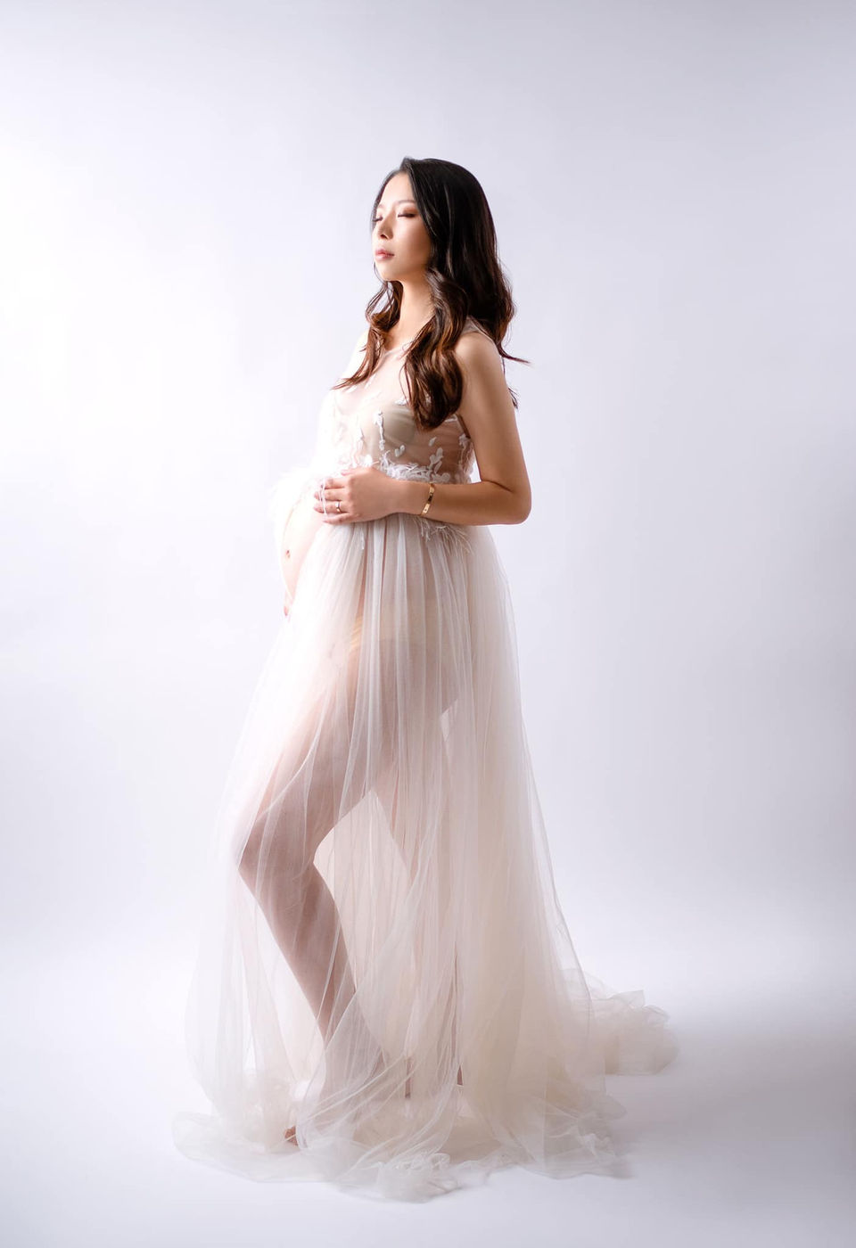stunning woman in sheer white maternity gown closes her eyes and holds her bump for a heavenly soft and elegant maternity portrait