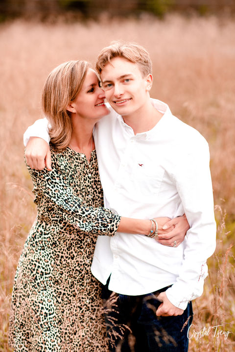 mom hugs and kisses son on the cheek for a mother son portrait standing in a tall, hay field.