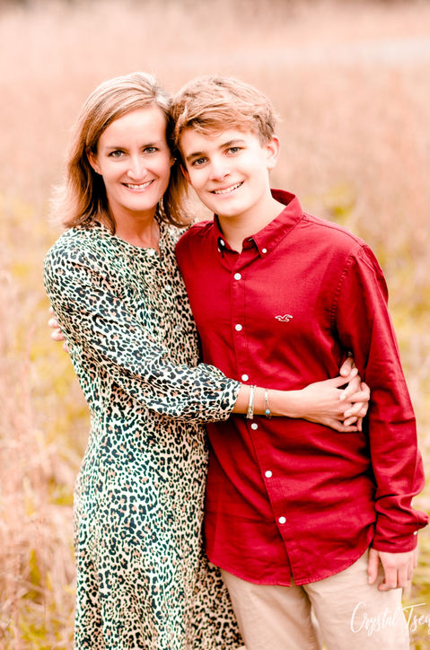 mom hugs her young son close as they stand in a tall, hay field.