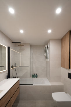 Baño moderno reformado con bañera, mobiliario de madera, espejo iluminado y ducha negra.