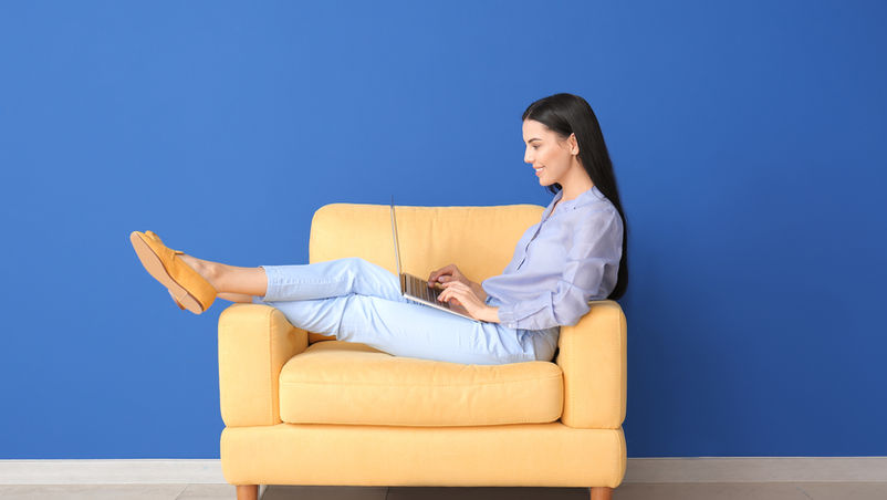 "A young woman wearing a light blue long sleeve shirt, seating on a sofa in a living room, on a laptop searching online for the fastest internet providers"