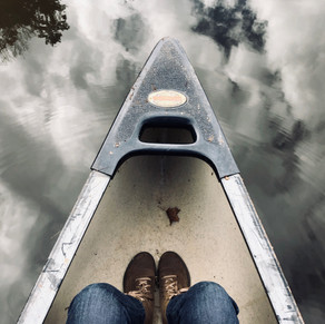 A birds eye view that captures the front end of a canoe where a persons feet can be seen. The canoe is gliding through still water which reflects the clouds above.
