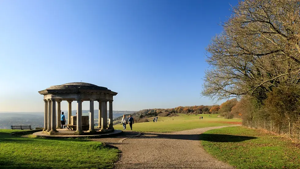 People walking near a stone monument on top of Reigate Hill on a sunny day, with a clear blue sky. Grass and bare trees surround the open landscape.