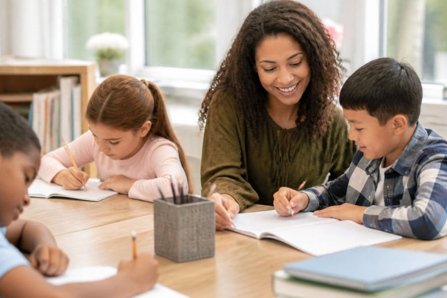 Children seated at a wooden table working on homework with a smiling adult staff member providing guidance in a bright, calm learning environment with natural light from windows