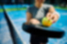 Frau hält einen gelben Padelball über einem Padelschläger auf einem blauen Indoor‑Court