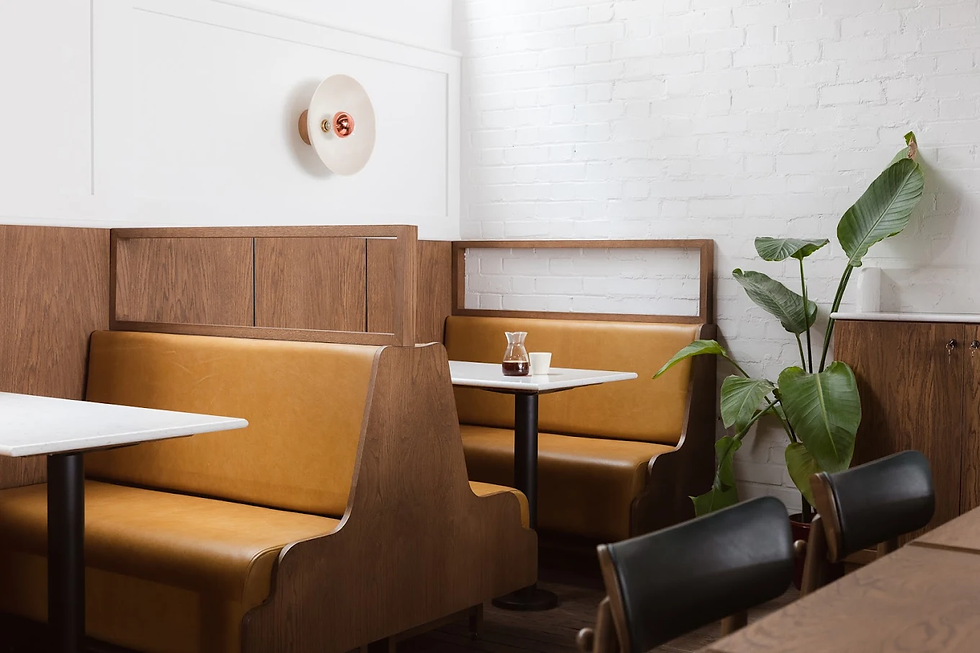 Cozy cafe interior with tan leather booths, white brick wall, round light fixture, and green plant. White table with coffee pot and cup.