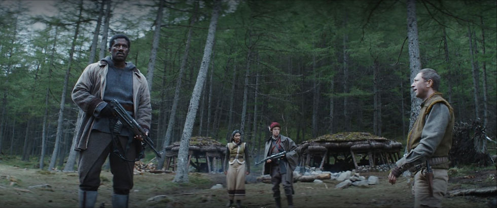 Four people stand in a forest clearing, three with rifles. Huts in the background, setting a tense, rustic scene.