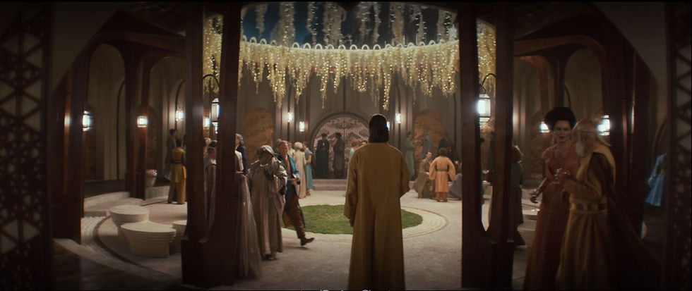 People in elegant attire gather in a lavish hall with ornate columns and a chandelier. Mood is formal and sociable.