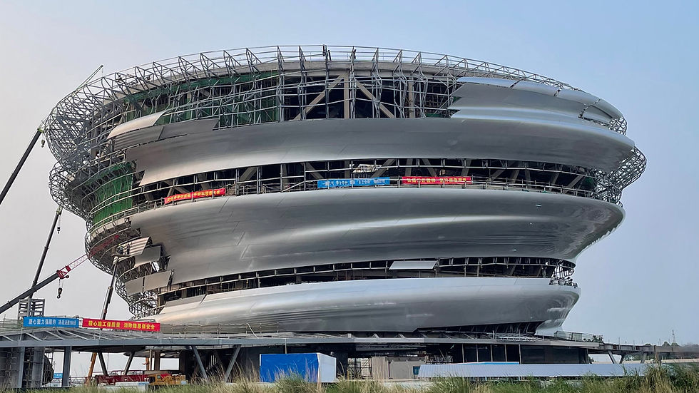 Futuristic, spiral-shaped building under construction with cranes around. Metallic exterior, blue sky, and red banners. Modern architecture.