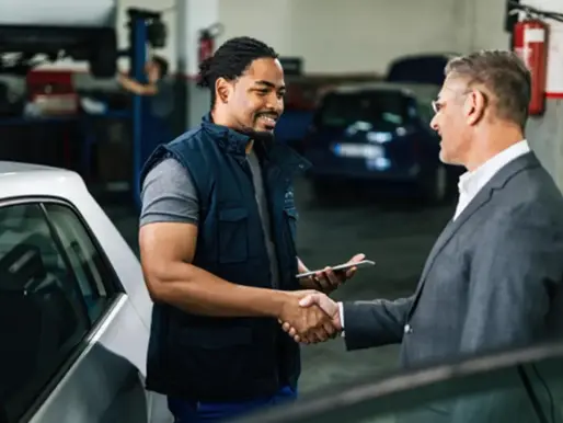 Inside an auto repair shop, a male customer is greeted warmly by an African‐American repairman with a big smile.
