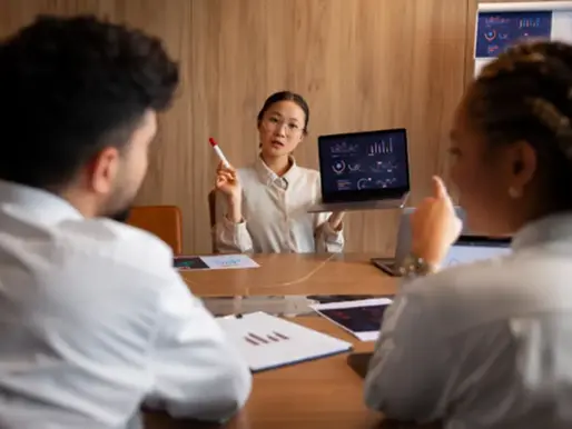 A group of office workers seated around a desk or conference table are looking at charts and graphs displayed.