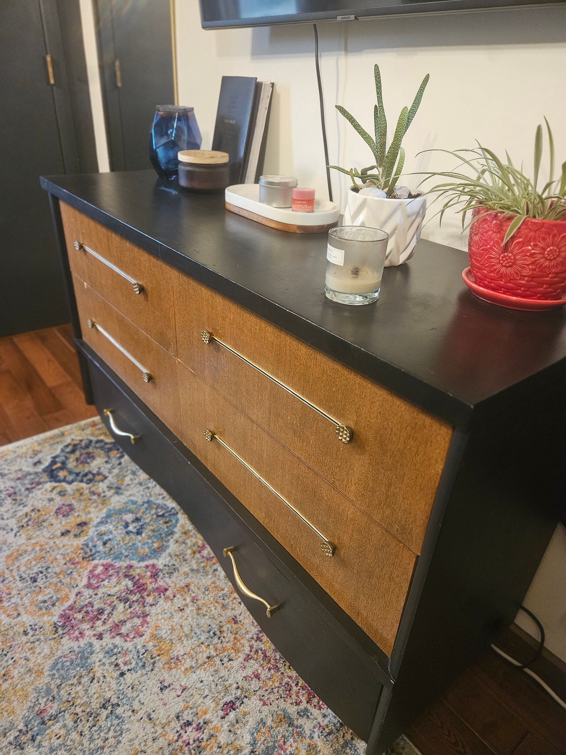 A side view of the lowboy dresser after makeover. The frame and the bottom 2 drawers have been painted a soft black. The top 1 drawers have been left in their original state with a golden veneer finish. The original brass hardware have been updated with a brighter gold paint. The dresser is accessorized with plants, candles and a lamp.