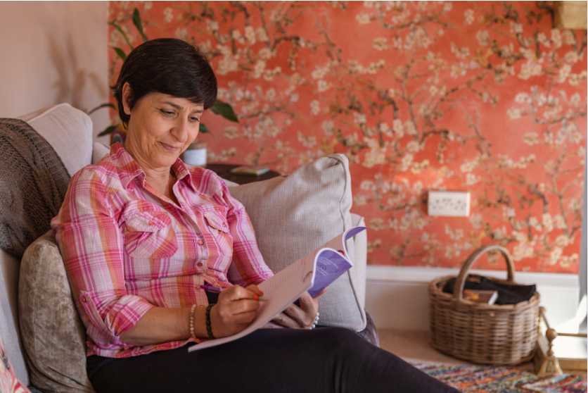 Carmela in a pink plaid shirt sits on a sofa, writing in a journal, with floral wallpaper and a wicker basket behind her.
