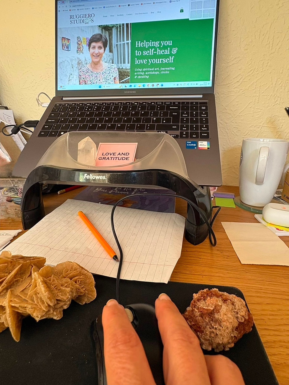 Home office desk with laptop on Ruggiero Studios site, hand on mouse, crystals and a Love and Gratitude note beside a mug.