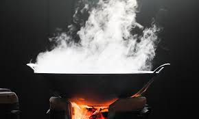 A black wok emitting steam sits on a lit stove against a dark background, creating a warm and dynamic atmosphere with orange flames.