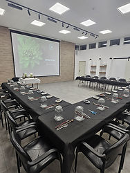 מרחב מוכן להעברת סדנה קבוצתית לבניית טרריום. השולחנות מסודרים בצורה יפה בצורת ח' מול מקרן שמקרין מצגת יפה. השולחנות ערוכים עם מפה שחורה, טרריום אישי וכלי עבודה אישיים לכל משתתף