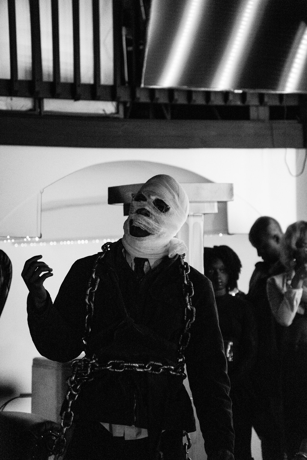 Man in bandages poses with chains in front of people, black and white.