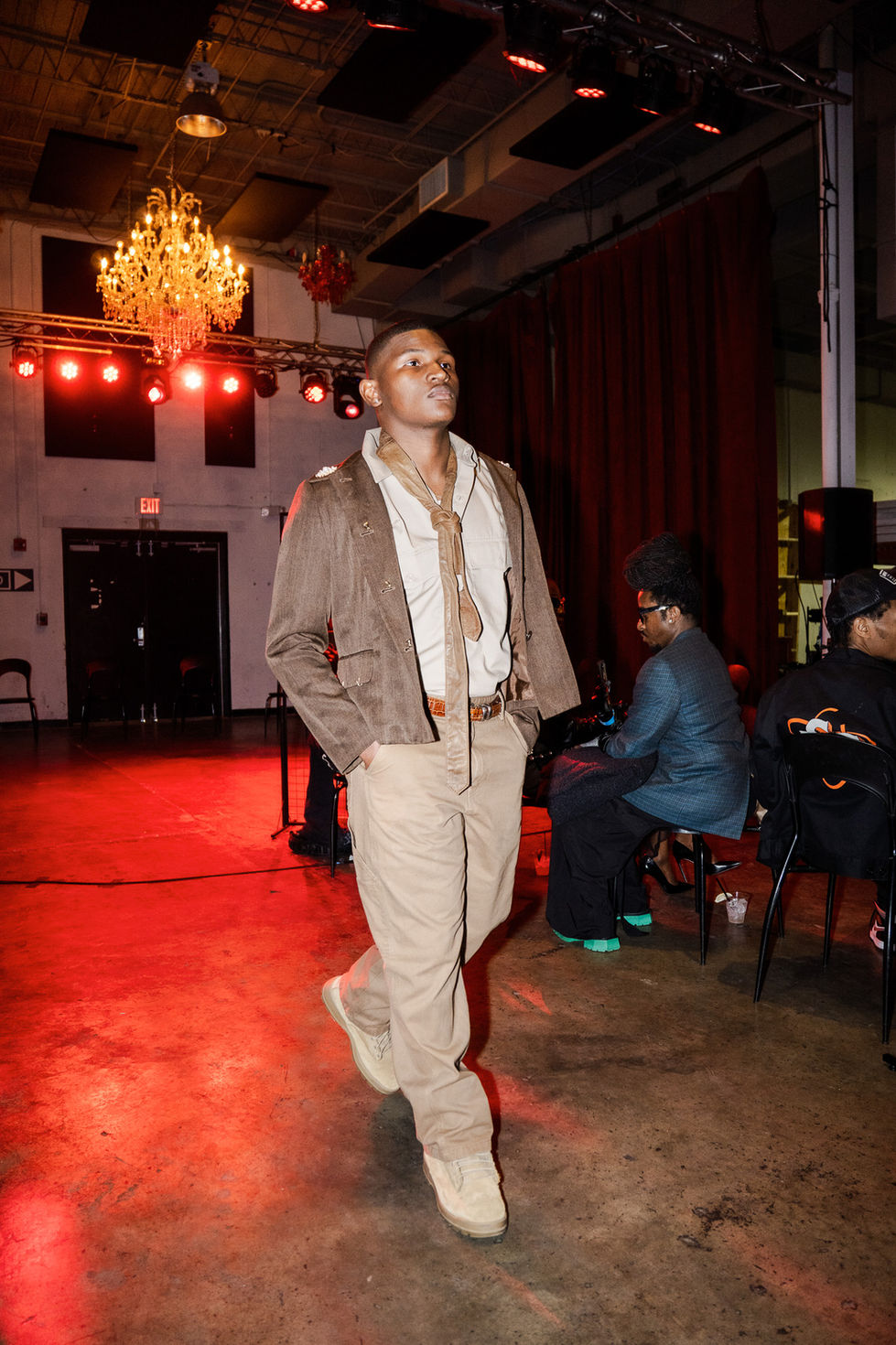 Male model in brown blazer and beige pants walks a fashion runway.