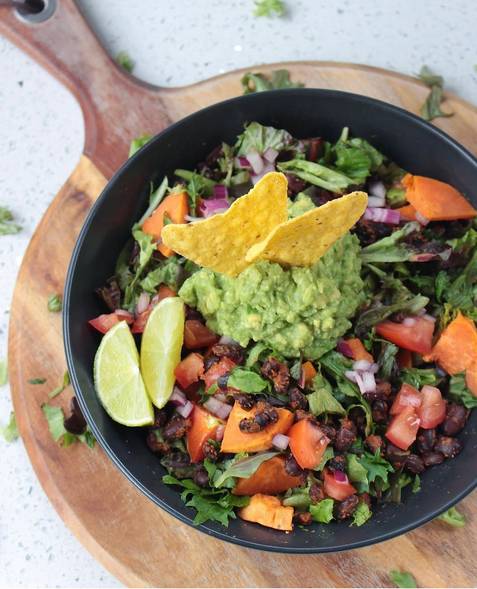 Black bowl of colorful salad with guacamole, tortilla chips, black beans, tomatoes, and lime on a wooden board