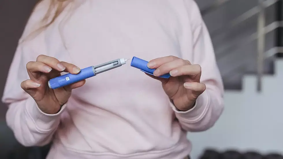 Person in pink sweater holds a blue insulin pen, unscrewing cap. Background shows blurred stairs. Focus on pen detail.