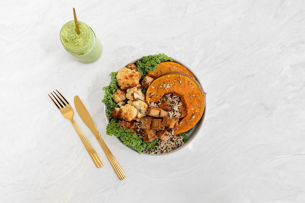 A bowl with roasted cauliflower, squash, rice, and kale on a marble surface. Gold cutlery and a jar of green smoothie are beside it.