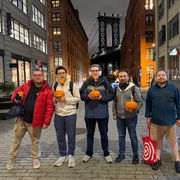 Five members stand holding carved pumpkins at night. The Manhattan bridge is in the background.