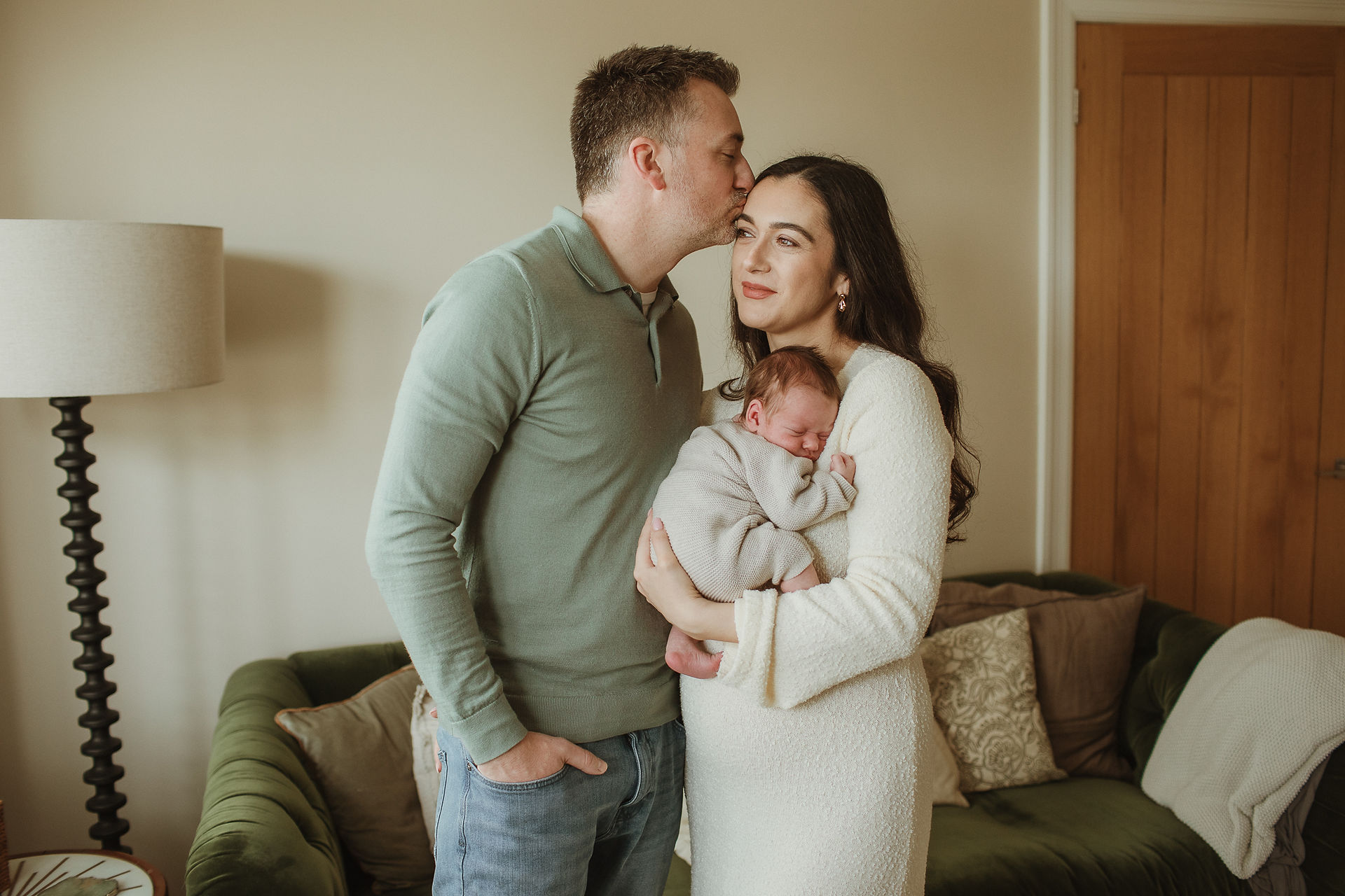 A colour image of mother holding a baby in a living room setting while dad stands next to her and kisses her on the head. Relaxed happy parents, taken during an in home newborn photoshoot in Chorlton.