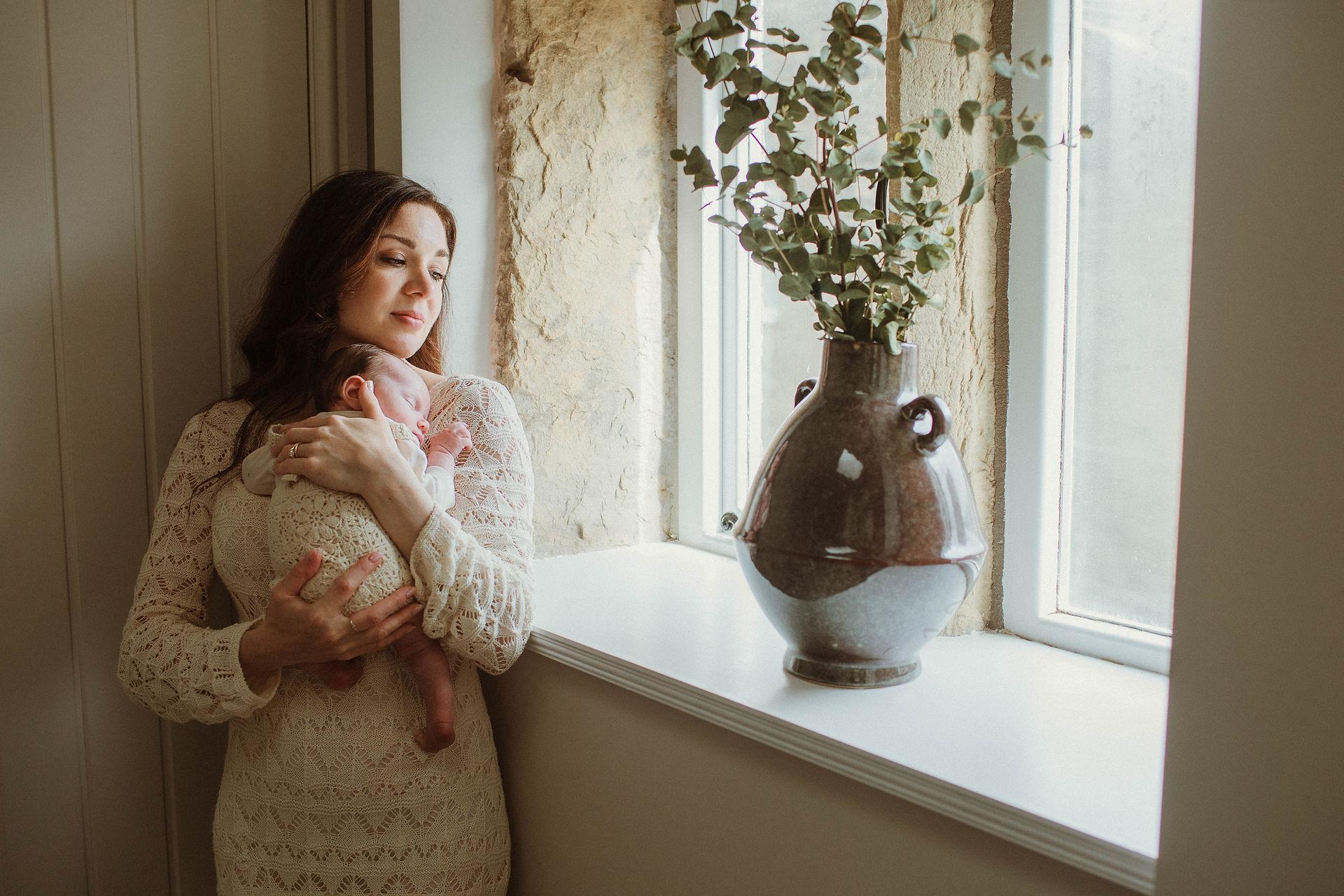 Mother holds her sleeping baby next to a window. Light floods in during a relaxed home newborn photoshoot in Manchester.
