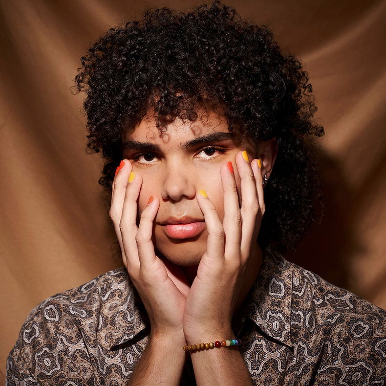 koblizzy poses up against a brown backdrop in a brown patterned shirt with their hands placed on either side of their face.