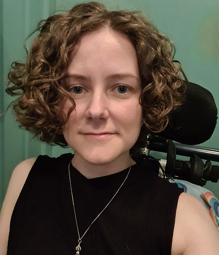 White woman with brown, chin length curly hair wearing a black top looks directly at the camera. wheelchair headrest in back