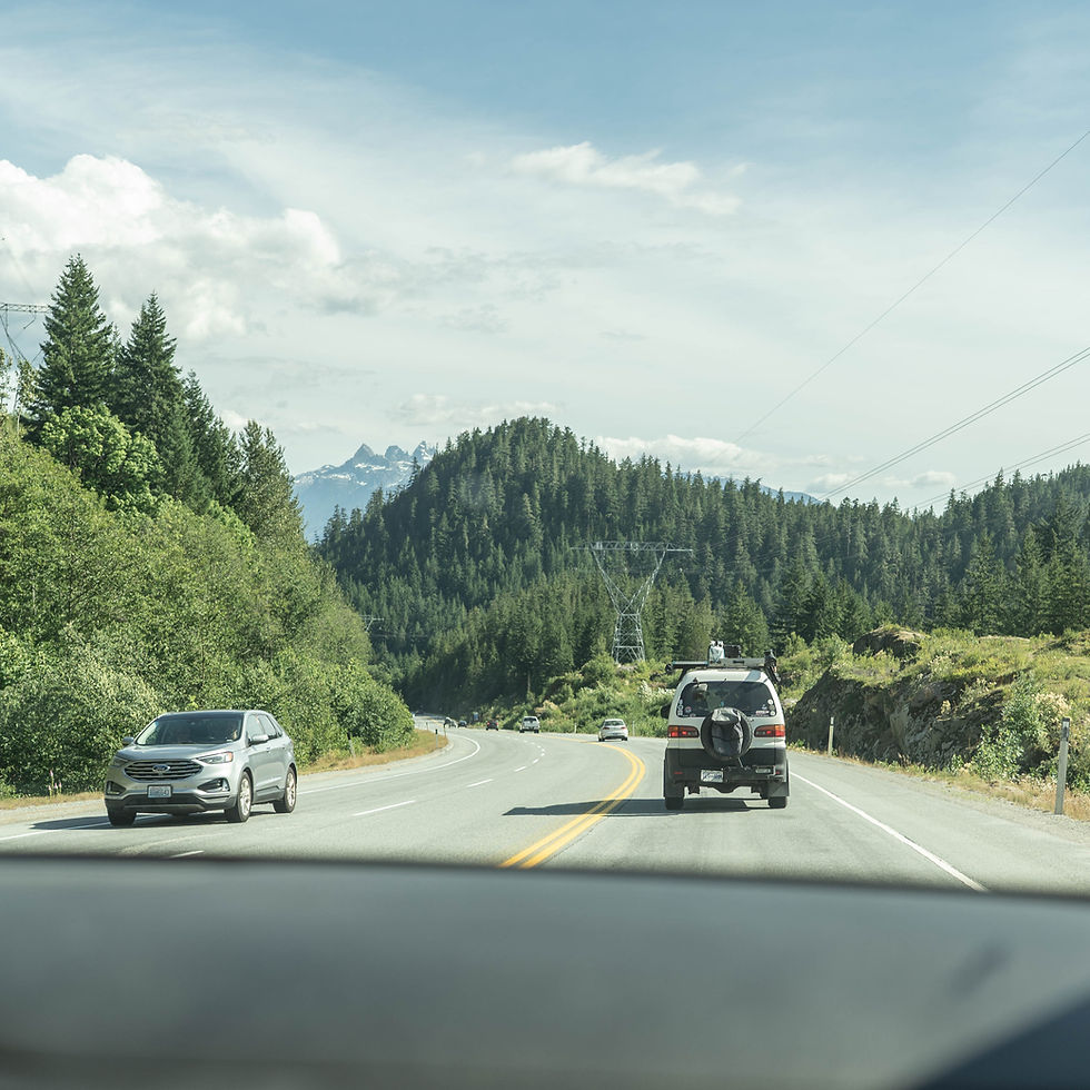 Cars drive on a scenic road surrounded by lush green trees and mountains. Overhead, power lines stretch across a partly cloudy blue sky.