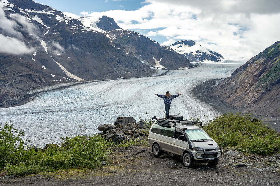 Person stands on van roof with arms outstretched, overlooking a vast glacier amid mountainous terrain, under a partly cloudy sky.