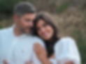 Woman wearing white dress and engagement ring smiles at the camera while holding the arm of her fiance