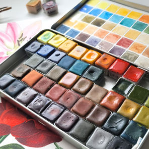 Close-up of a watercolor paint palette with handmade paints in various colors, placed on a botanical watercolor painting in an artist’s studio.