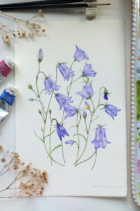 A delicate watercolor painting of multiple purple bell-shaped flowers with green stems, set on white paper with light pencil sketches. Surrounded by art supplies including paintbrushes, a watercolor palette with mixed colors, and dried plant stems, showcasing a serene floral artwork.