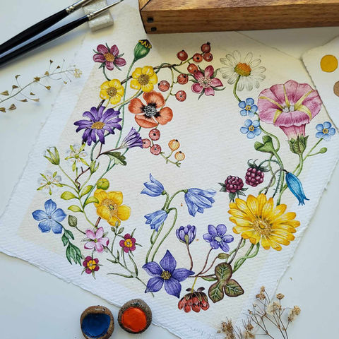 A stunning watercolor painting showcasing a vibrant circular arrangement of colorful flowers, including purple, yellow, pink, orange, and blue blooms, with green leaves, berries, and delicate stems on textured white paper. Surrounded by art supplies like paintbrushes, watercolor paints, and dried plants, this eye-catching floral artwork is perfect for art lovers and nature enthusiasts.