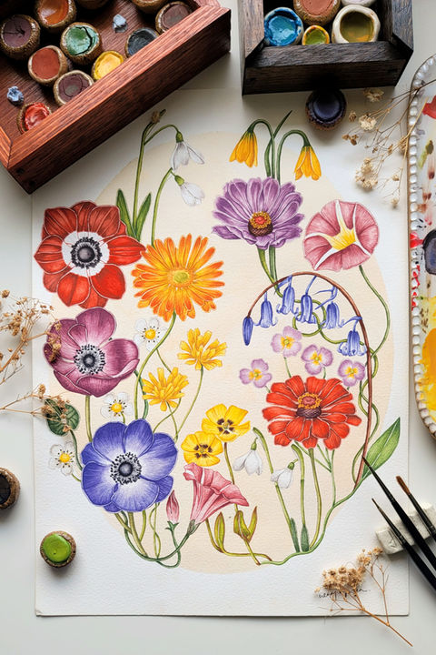 A stunning watercolor painting showcasing a vibrant circular arrangement of colorful flowers, including purple, yellow, pink, orange, and blue blooms, with green leaves, berries, and delicate stems on textured white paper. Surrounded by art supplies like paintbrushes, watercolor paints, and dried plants, this eye-catching floral artwork is perfect for art lovers and nature enthusiasts.