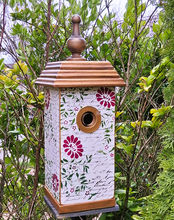 A column style birdhouse with a finial rooftop, painted white with pink flowers and a script font background